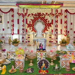 A beautiful Seemantha setup with a red and white floral backdrop. The artificial grass flooring and detailed doll arrangements in the foreground add a touch of charm and tradition to the event.