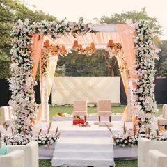 A beautiful pastel-themed mandap, with pillars heavily adorned with white and pink roses. The soft peach draping and hanging floral chandeliers create a dreamy and romantic atmosphere for an outdoor ceremony.