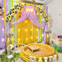 I design classic Haldi ceremony spaces perfect for home functions. This setup features a beautiful floral arch with sunflowers over a yellow and lavender draped backdrop, with a traditional brass-style urli for seating and fun props to get the party started.