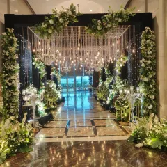 A glamorous and sparkling entrance tunnel. This structure combines a ceiling of hanging crystals and lights with pillars of white flowers, creating a dazzling welcome for a grand reception.