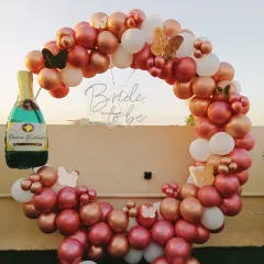 This outdoor bridal shower setup features a rose gold and white balloon arch against the open sky. The design includes butterfly accents and a champagne foil balloon, creating a beautiful photo opportunity for the bride-to-be.