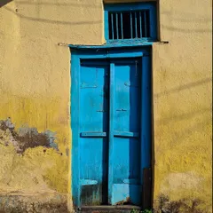 A vibrant blue door provides a striking contrast against a weathered yellow wall. This simple yet powerful composition highlights the bold use of color in traditional Indian village architecture.