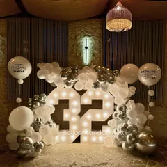 Make your milestone shine. This 32nd birthday setup features large marquee light numbers, framed by a chic white and chrome silver balloon garland, creating a sophisticated and celebratory focal point.