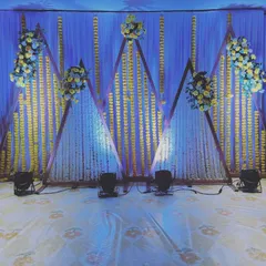 A modern and geometric stage design for a Sangeet or reception. The backdrop features triangular frames decorated with white and yellow flower strings.