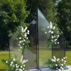 This modern and elegant decor piece, featuring mirrored panels adorned with white flowers and fresh greenery, serves as a beautiful photo backdrop for an outdoor event. It is a perfect blend of nature and contemporary design.