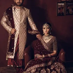 This is one of our classic bridal couple sets, perfect for the main wedding ceremony. The bride is wearing a deep maroon velvet lehenga with detailed zardozi work, and the groom is in a cream and gold patterned sherwani with a matching maroon stole. We help you get this complete, perfectly coordinated look without the stress.