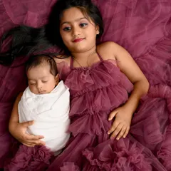 A sister's love is a special gift. This overhead view captures a tender moment between a proud big sister and her sleeping newborn sibling, both nestled in a beautiful cascade of fabric.