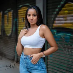 A classic urban street style portrait. The combination of a simple white crop top, denim jeans, and a graffiti-covered shutter in the background gives this shot a raw and confident feel.