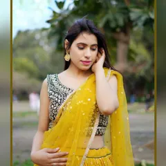 A soft and elegant outdoor fashion shot. The model, dressed in a yellow lehenga with mirror work, is captured in a gentle, contemplative pose against a natural park background.