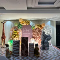 A sophisticated jungle-themed first birthday stage. This setup features large animal props like a giraffe and elephant, animal-print pedestals, and a custom backdrop with the child's name.
