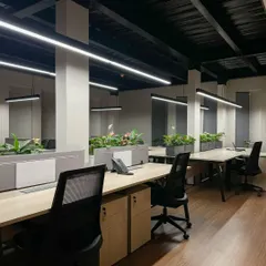 A view across the open-plan work floor, featuring rows of workstations with integrated planters. The industrial-inspired exposed ceiling is fitted with modern linear lighting to ensure a bright and energizing environment.