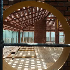 A view through a circular opening in a brick wall during construction. This shot highlights the beautiful play of light and shadow created by a latticed roof, turning a simple material into a dynamic architectural element.