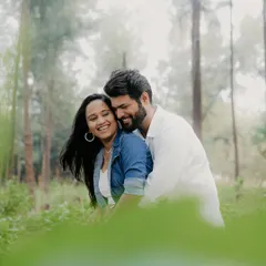 A couple shares a hug in a sun-dappled forest, framed by green leaves in the foreground. This composition creates a sense of intimacy and being lost in their own world.