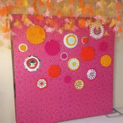 A creative and colorful photo wall for a mehendi or sangeet. The pink patterned backdrop is decorated with circular patterns made from marigold flowers and mirrors.