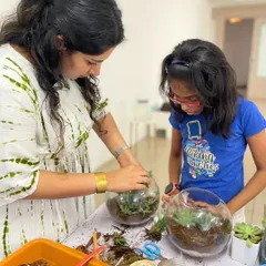 An instructor helps a young girl arrange the plants in her round glass bowl terrarium, teaching her about placement and care.