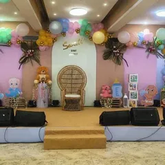 A full stage decoration we did for a large baby shower event. The setup includes a central wicker chair for the mom-to-be, flanked by teddy bear and baby doll props, all set against a pastel pink and blue backdrop.