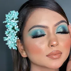 A detailed close-up of a blue smokey eye with a touch of glitter on the lid. This image shows the precise blending and shimmer application, complemented by a soft, peachy blush and a glossy lip.