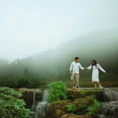 Walking hand in hand across a natural stone bridge over a smaller, gentle cascade. The misty forest background adds to the romantic, ethereal atmosphere of this destination shoot.