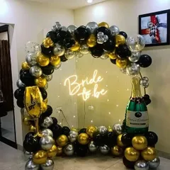 A classic black, gold, and silver themed bachelorette party photo booth. The balloon arch is accented with large foil balloons shaped like a champagne bottle and a wine glass, adding a celebratory touch to the "Bride to be" neon sign.