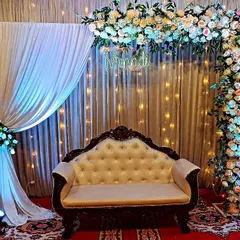 An elegant Mehndi stage with a floral arch, a "Mehndi" neon sign, and shimmering curtains with fairy lights. The traditional sofa and side vases add a touch of class to the setup.