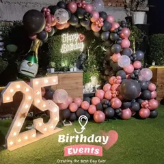 A stylish outdoor setup for a 25th birthday on a lawn. The decor features a pink and black balloon arch, a neon sign, and large marquee numbers.