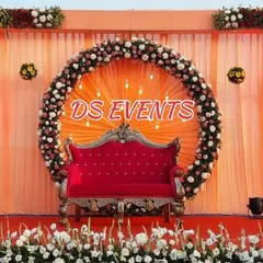 This reception stage features a bold red sofa set against a peach-draped backdrop. The circular floral arrangement is accented with warm bulb lights, adding a touch of glamour to the outdoor setup.