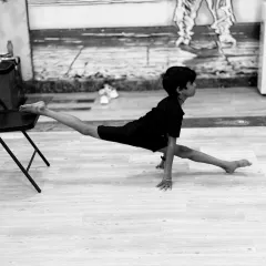 A beautiful black and white shot of a young dancer practicing a split using a chair for support. This demonstrates our focus on technique and safe progression.