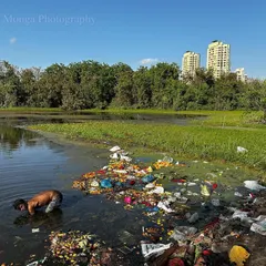 A young boy wades in a waterway choked with plastic and garbage, with city buildings in the background. The debris of our wasteful lifestyles intrudes on every natural space.