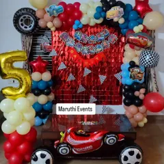 A high-speed 'Cars' themed party for a 5th birthday. The decoration includes a red shimmer backdrop, a black and red balloon arch, and a large race car foil balloon.