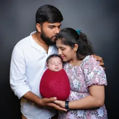 A classic family portrait featuring parents holding their newborn, who is wrapped in a vibrant red swaddle. The simple black background makes the family the center of attention.