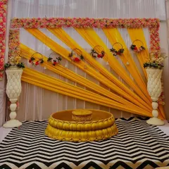 A simple yet elegant Haldi setup. The backdrop uses yellow drapes arranged in a sun-ray pattern, framed by a delicate floral border. The patterned rug adds a modern touch to the otherwise traditional decor.
