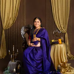 A regal portrait of an expecting mother seated on an ornate chair. She is wearing a rich purple silk saree, smiling warmly in a traditionally styled studio setting.