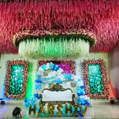 This grand stage was designed for a 'Welcome Baby' ceremony, featuring an ornate white throne-style sofa for the family. The backdrop includes a dense balloon arch in shades of blue and white, custom banners, and large gold foil balloons spelling out the baby's name, all under an elaborate ceiling of hanging flowers.