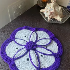 A beautiful crochet table mat with a purple and white floral petal design, adding an elegant and handmade accent to a side table.