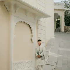 A solo portrait of the groom looking sharp in his traditional attire against the pristine white architecture of a Udaipur palace. It's a moment of quiet confidence before the big day.