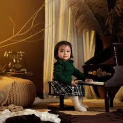A little girl in a green sweater and plaid skirt sits at a miniature piano in a cozy, rustic-themed studio setup.
