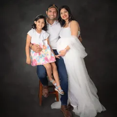 A classic and happy family portrait. The mom-to-be, dressed in a beautiful white gown, is surrounded by her loving husband and daughter.