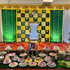 A traditional green and yellow themed Seemantham decor. The stage includes a floral backdrop, a ceremonial throne, and a wide array of beautifully arranged thamboolam plates.