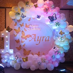 A whimsical butterfly theme for a first birthday party. The circular backdrop is adorned with golden butterflies and a pastel balloon garland, complemented by a light-up number '1'.