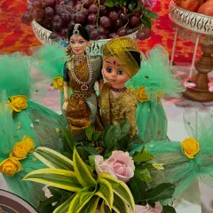 An adorable couple doll arrangement perfect for an engagement or wedding. The dolls are dressed in traditional attire and placed amidst fresh flowers and a platter of grapes.