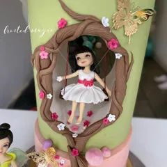 This fairy-themed cake features a little girl on a swing, framed by tree branches. All the figures are handmade to create a unique, storybook feel.