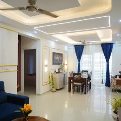 This wide view showcases a cohesive living and dining area, defined by its elegant false ceiling with recessed and profile lighting. The combination of a plush blue sofa and a classic wooden dining set creates distinct zones within the open-plan space.