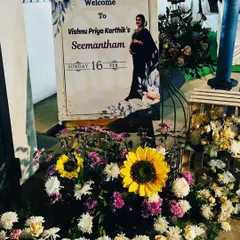 A personalized welcome board for a Seemantham ceremony, featuring a photo of the mother-to-be. The board is surrounded by an arrangement of sunflowers and other colorful flowers.