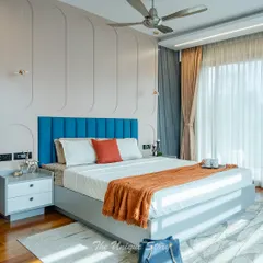 A bright and airy master bedroom featuring a bold blue channel-tufted headboard. The wall behind is adorned with curved pastel paneling, adding a soft, architectural element. The room is designed with warm wood flooring and large windows for abundant natural light.