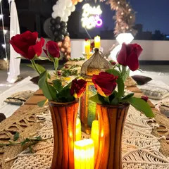 A close-up detail of the tablescape from a romantic terrace dinner. The decor includes red roses, candle-lit lanterns, and a macrame table runner, creating a warm and intimate ambiance.