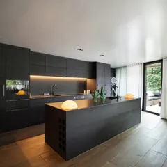 This modern pantry and breakout space was created for a corporate office, featuring sleek, handleless cabinetry and a large central island. Warm, under-cabinet lighting and natural wood floors balance the dark, minimalist aesthetic, providing a welcoming area for employees to connect.