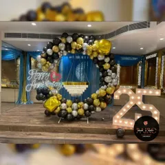 A luxurious 25th-anniversary ring backdrop with black, gold, and silver balloons. The setup is accented with a chandelier centerpiece and large "25" marquee numbers.