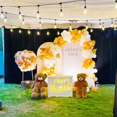 An adorable teddy bear themed birthday party set up outdoors on a lawn. The decor features two large teddy bears, a neon "Happy Birthday" sign, and an elegant balloon arch in white and orange.