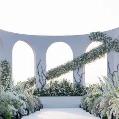 A direct view of the altar, featuring a clean, curved wall with modern arches. A sweeping garland of white flowers cuts across the structure, adding a dynamic and artistic element to the classic white wedding theme.