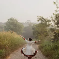 A wide shot capturing the dancer's joyful expression of freedom. With arms outstretched on a country road, the movement of her anarkali and the misty background create a sense of boundless energy.